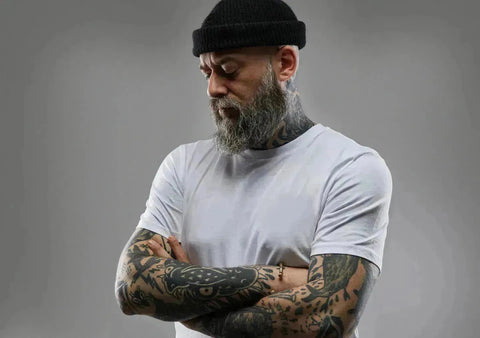 Bearded man with tattoos in a white t-shirt and black cap from Hukone collection.