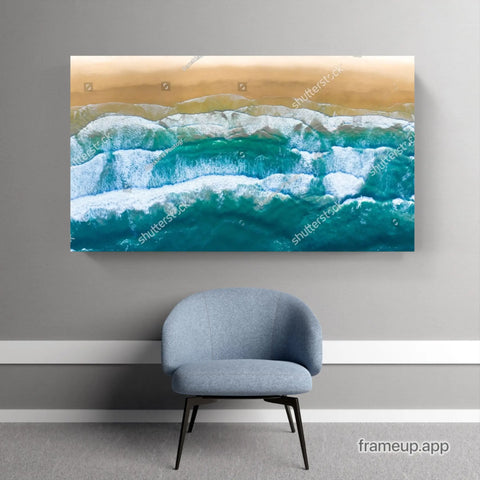 Aerial beach photograph showing turquoise waves breaking on sandy shore mounted as wall art above a gray accent chair.